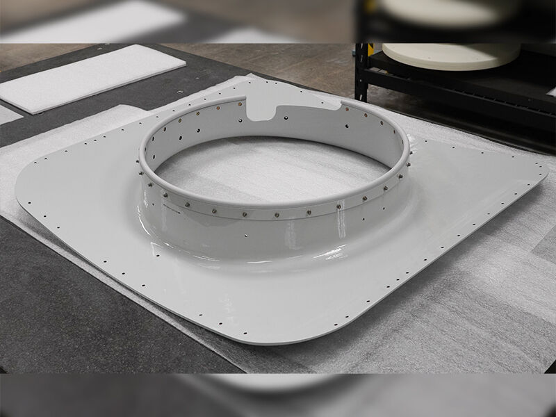 Fairing aerostructure on a table in a manufacturing environment. Curved white shape with drill holes along the edges and metallic inserts around the central hole.